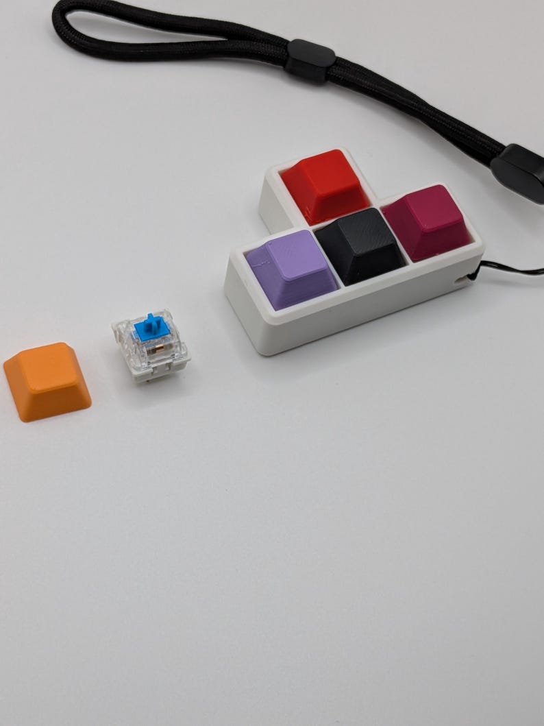 May include: A collection of colorful mechanical keyboard keycaps in red, black, purple, and magenta, arranged in a white holder. An orange keycap and a clear switch are also visible. A black wrist strap is in the background.