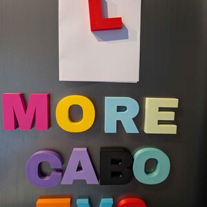 May include: Colorful magnetic letters spelling "MORE CABO TVS" on a gray surface. A red "L" is attached to a white paper. The letters are in various colors, including pink, yellow, purple, black, blue, and orange.