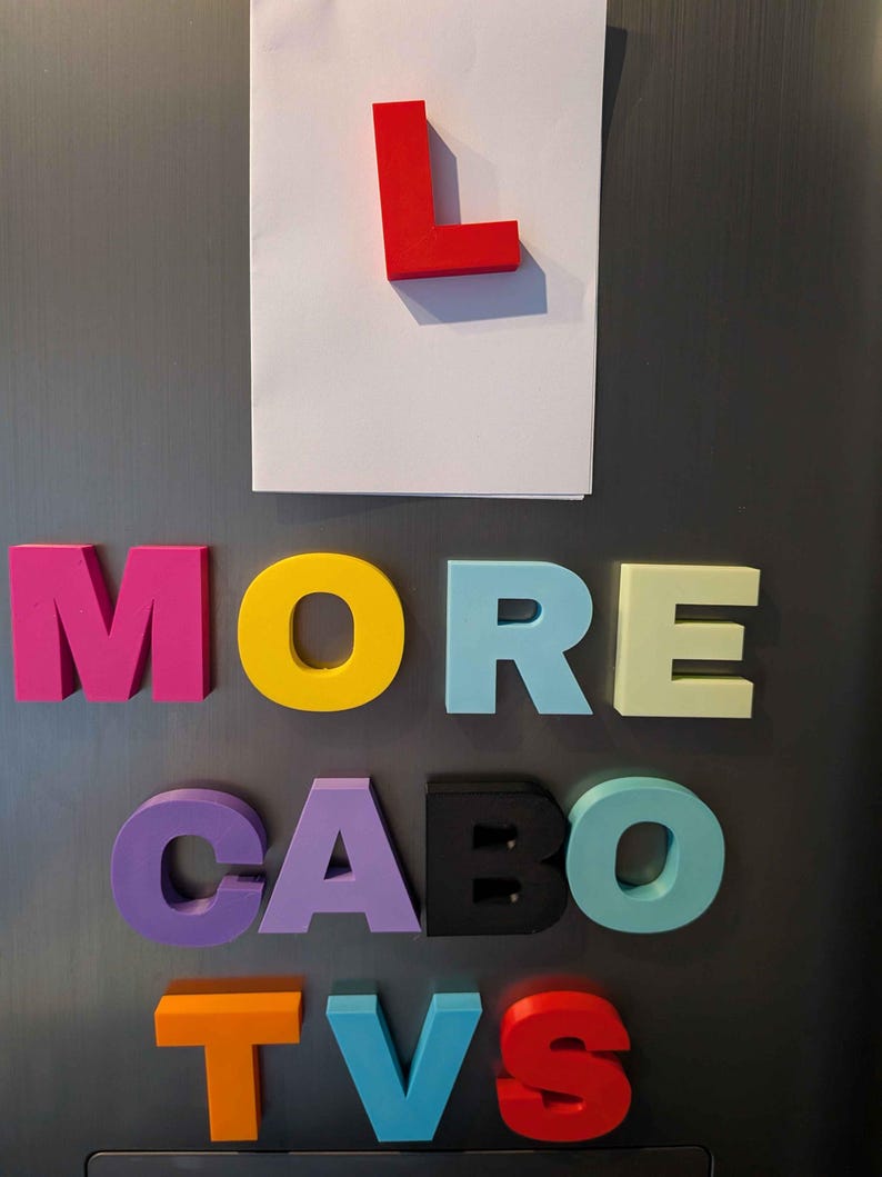 May include: Colorful magnetic letters spelling "MORE CABO TVS" on a gray surface. A red "L" is attached to a white paper. The letters are in various colors, including pink, yellow, purple, black, blue, and orange.