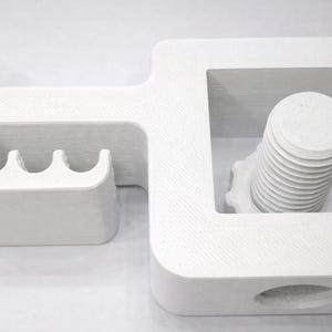 Puede incluir: Un objeto geom&eacute;trico blanco con un dise&ntilde;o en forma de C, que presenta un perno y una serie de ranuras curvas. El objeto parece estar impreso en 3D, con l&iacute;neas de capa visibles. El dise&ntilde;o incluye un perno cil&iacute;ndrico con un v&aacute;stago roscado.
