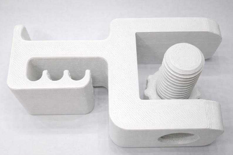 Puede incluir: Un objeto geom&eacute;trico blanco con un dise&ntilde;o en forma de C, que presenta un perno y una serie de ranuras curvas. El objeto parece estar impreso en 3D, con l&iacute;neas de capa visibles. El dise&ntilde;o incluye un perno cil&iacute;ndrico con un v&aacute;stago roscado.