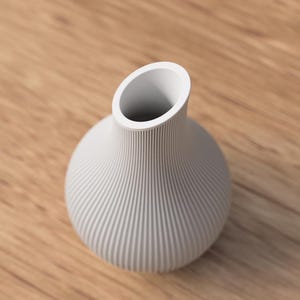 May include: A white, ribbed ceramic vase with a rounded body and an angled opening. The vase sits on a light brown wooden surface. The vase's design features vertical grooves, adding texture to its smooth, matte finish. This decorative vase is suitable for floral arrangements.