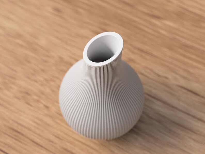 May include: A white, ribbed ceramic vase with a rounded body and an angled opening. The vase sits on a light brown wooden surface. The vase's design features vertical grooves, adding texture to its smooth, matte finish. This decorative vase is suitable for floral arrangements.
