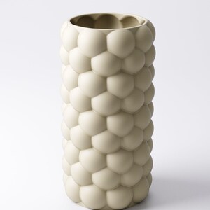 May include: A tall, off-white vase with a textured surface of rounded, bubble-like shapes. The vase has a cylindrical form and a wide opening at the top. The design gives the vase a unique, modern aesthetic. The vase is suitable for home decor.