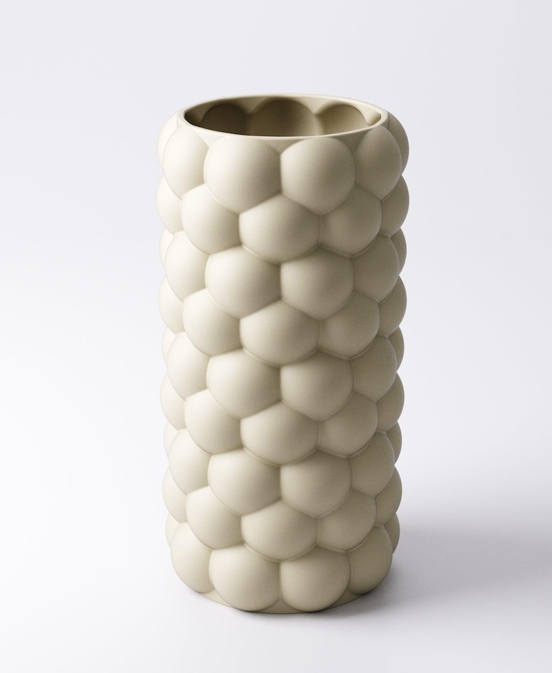 May include: A tall, off-white vase with a textured surface of rounded, bubble-like shapes. The vase has a cylindrical form and a wide opening at the top. The design gives the vase a unique, modern aesthetic. The vase is suitable for home decor.