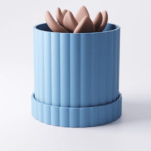 May include: A blue cylindrical planter with vertical ridges, holding a succulent plant. The plant has multiple fleshy, petal-like leaves in a muted pink color. The planter sits on a matching blue saucer.