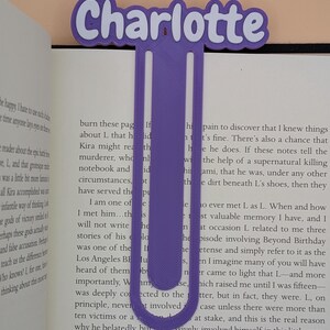 May include: A purple 3D-printed bookmark shaped like a large paperclip with the name Charlotte displayed in a bold, white-outlined font at the top. The bookmark is positioned vertically between the pages of an open book with visible text.