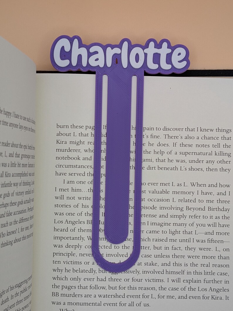 May include: A purple 3D-printed bookmark shaped like a large paperclip with the name Charlotte displayed in a bold, white-outlined font at the top. The bookmark is positioned vertically between the pages of an open book with visible text.