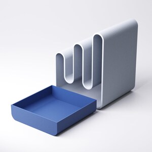 May include: A modern desk organizer in shades of gray and blue. The organizer features a wavy, vertical design with a built-in tray. The tray is a solid blue, while the main structure is gray with white accents. A stylish and functional office accessory.