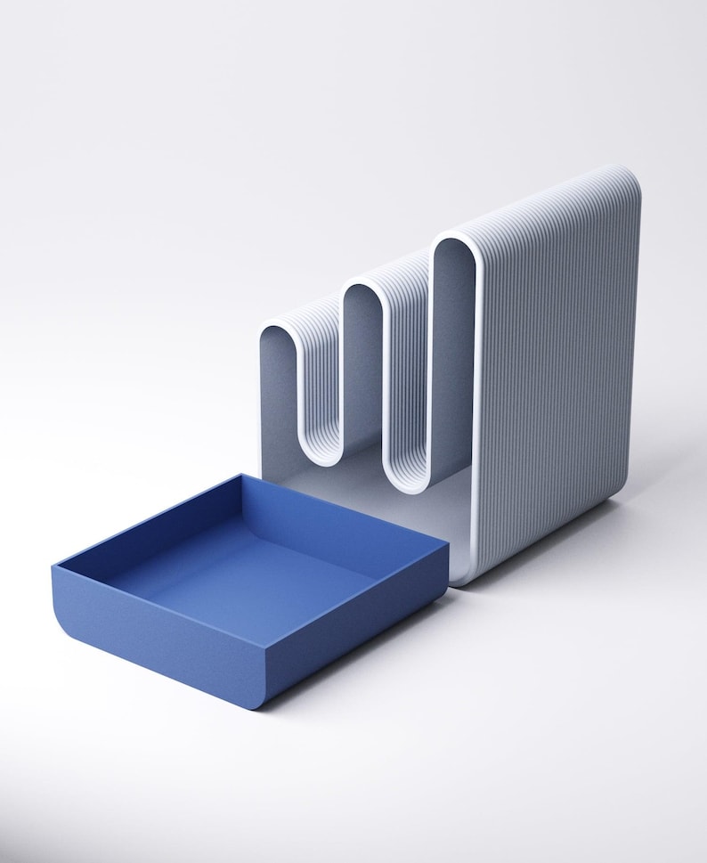 May include: A modern desk organizer in shades of gray and blue. The organizer features a wavy, vertical design with a built-in tray. The tray is a solid blue, while the main structure is gray with white accents. A stylish and functional office accessory.