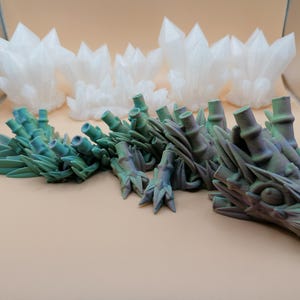 May include: A detailed 3D-printed dragon sculpture in shades of green and purple, with a textured surface. The dragon is positioned in front of several white, crystal-like structures. The objects are set against a neutral background.