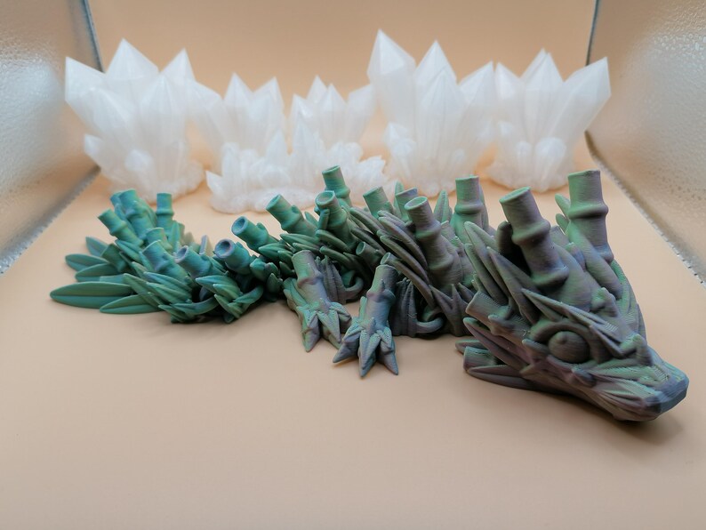 May include: A detailed 3D-printed dragon sculpture in shades of green and purple, with a textured surface. The dragon is positioned in front of several white, crystal-like structures. The objects are set against a neutral background.