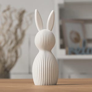 K&ouml;nnte beinhalten: Eine cremefarbene, dekorative Hasenfigur mit vertikalen Streifen. Der Hase hat lange, aufrechte Ohren und einen abgerundeten K&ouml;rper. Die Figur besteht aus einem strukturierten Material und steht auf einer Holzoberfl&auml;che. Der Hintergrund ist unscharf.