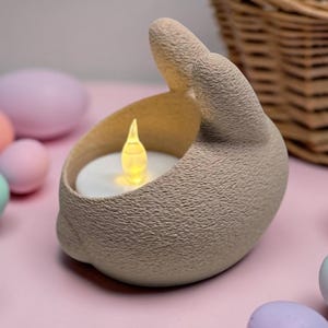 K&ouml;nnte beinhalten: Ein beigefarbener Hasen-Kerzenhalter mit einer flackernden LED-Kerze im Inneren. Ostereier in Pastellfarben umgeben den Kerzenhalter auf einer rosa Oberfl&auml;che. Ein Weidenkorb befindet sich im Hintergrund.