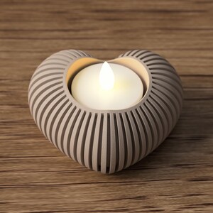 May include: A heart-shaped candle holder with a lit candle inside. The holder is light gray with vertical grooves, and the candle emits a warm, inviting glow. The holder sits on a wooden surface.