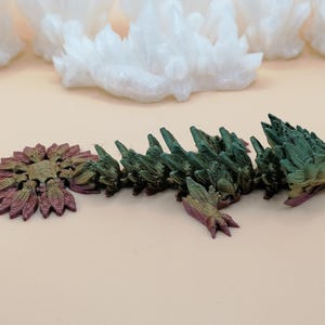 May include: A multi-colored, articulated dragon keychain. The dragon is primarily green with hints of purple and gold. The keychain is made of a 3D-printed material and has a silver-colored keyring.