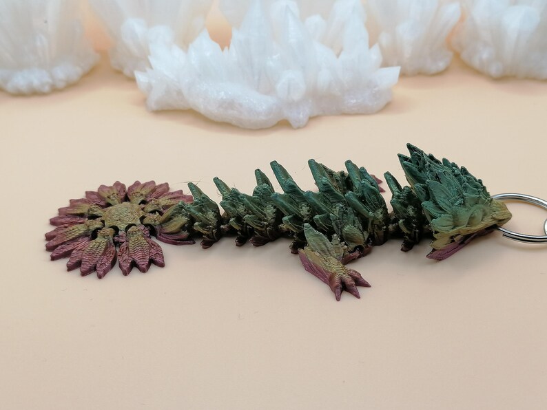 May include: A multi-colored, articulated dragon keychain. The dragon is primarily green with hints of purple and gold. The keychain is made of a 3D-printed material and has a silver-colored keyring.