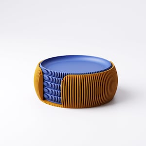 May include: A stack of blue plates inside a mustard yellow holder. The holder has a ribbed design. The plates are stacked neatly, with the top plate visible. The holder is a rounded shape, with vertical lines. The background is white.