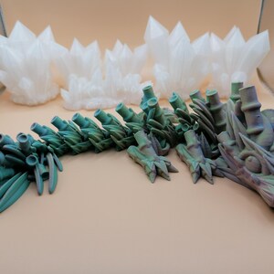 May include: A 3D-printed articulated dragon in shades of teal and purple, with a detailed, textured design. The dragon is posed on a neutral surface, with several white, crystalline formations in the background.
