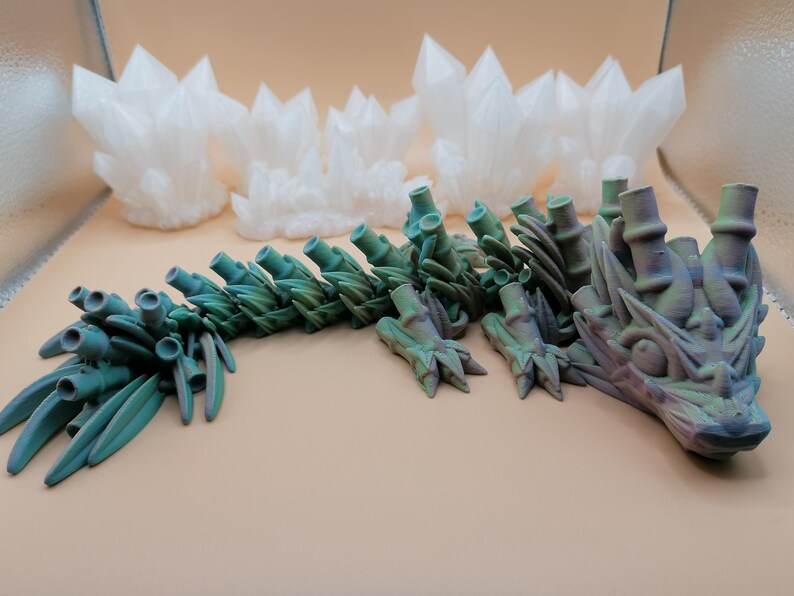May include: A 3D-printed articulated dragon in shades of teal and purple, with a detailed, textured design. The dragon is posed on a neutral surface, with several white, crystalline formations in the background.