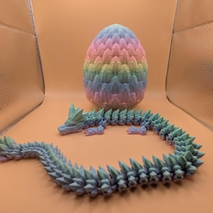 May include: A 3D-printed dragon and egg set. The dragon is a light blue-green color with a spiky design. The egg is a pastel rainbow of pink, yellow, blue, and green. The dragon is curled around the egg.
