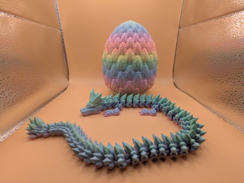 May include: A 3D-printed dragon and egg set. The dragon is a light blue-green color with a spiky design. The egg is a pastel rainbow of pink, yellow, blue, and green. The dragon is curled around the egg.
