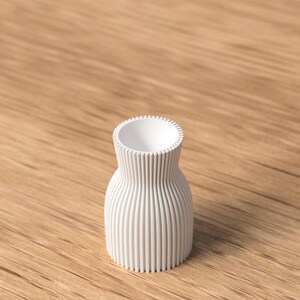 Pu&ograve; includere: Un piccolo vaso bianco con esterno a coste. Il vaso ha un corpo arrotondato e un'apertura stretta e strutturata. Il vaso &egrave; appoggiato su una superficie di legno.