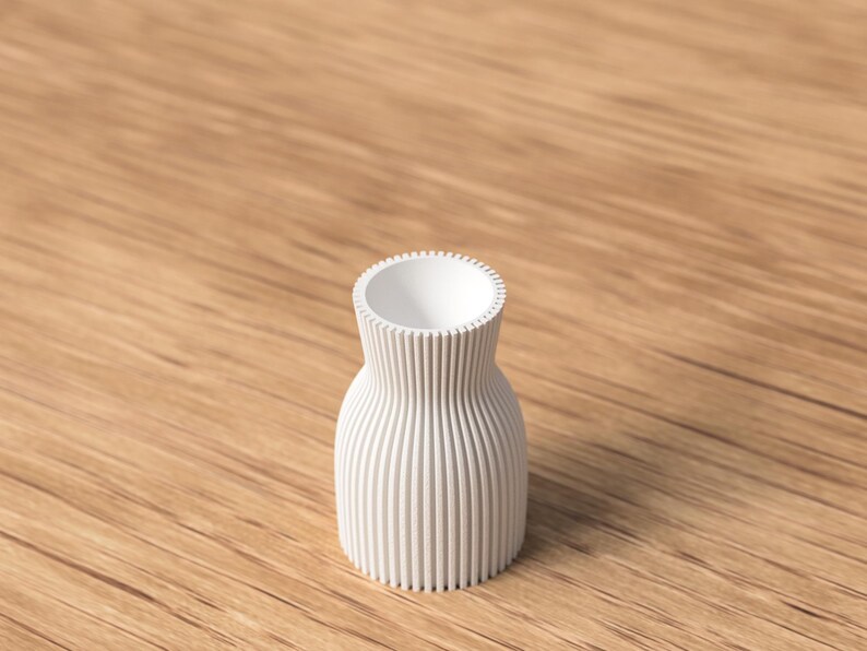 Pu&ograve; includere: Un piccolo vaso bianco con esterno a coste. Il vaso ha un corpo arrotondato e un'apertura stretta e strutturata. Il vaso &egrave; appoggiato su una superficie di legno.