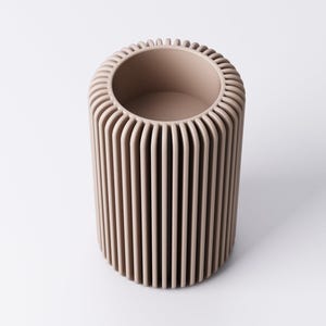 May include: A cylindrical, beige-colored vase with vertical ribbed detailing. The top rim has a rounded, textured edge. The interior is a smooth, open space. The vase is a neutral tone, suitable for various decor styles.