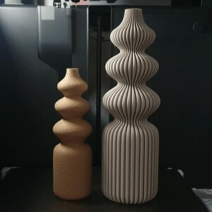 May include: Two decorative vases. One is a light brown, textured, gourd-shaped vase. The other is a tall, beige vase with vertical ribbed detailing. Both vases have a bulbous shape and are placed on a dark surface.