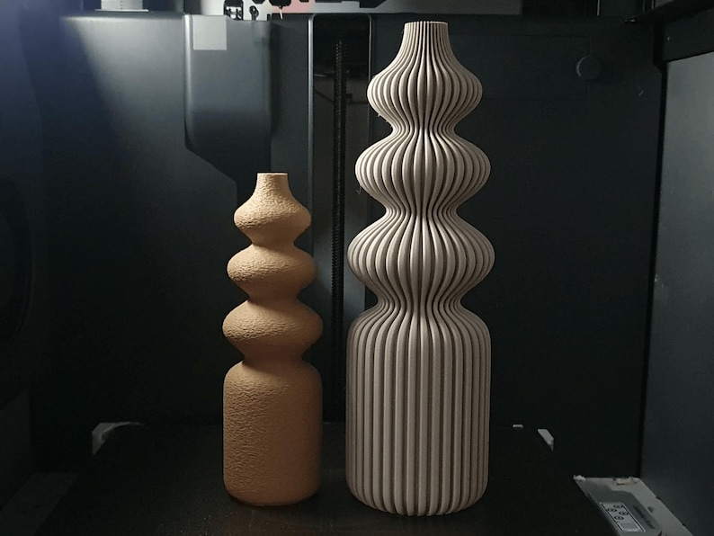 May include: Two decorative vases. One is a light brown, textured, gourd-shaped vase. The other is a tall, beige vase with vertical ribbed detailing. Both vases have a bulbous shape and are placed on a dark surface.