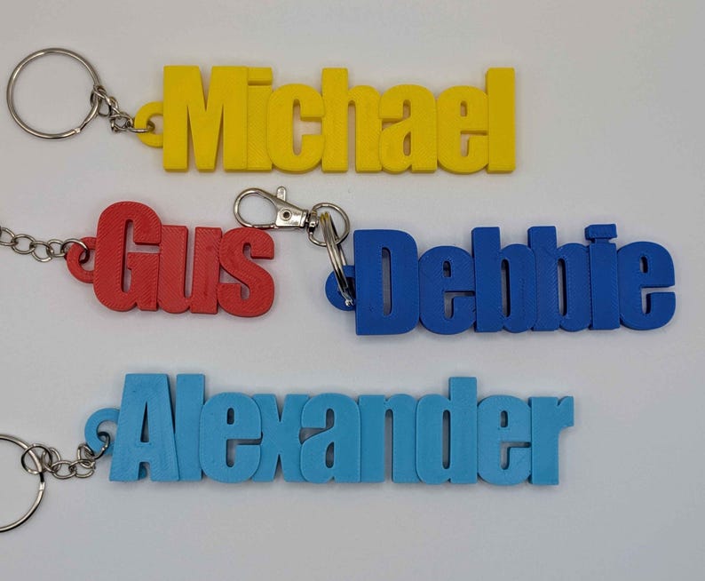 May include: Four colorful name keychains: yellow "Michael", red "Gus", blue "Debbie", and light blue "Alexander". Each keychain features a name in bold, block letters, attached to a metal keyring or clasp.
