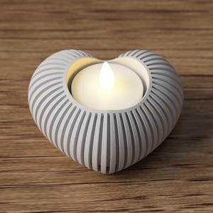 May include: A heart-shaped candle holder with a white, ribbed design. The holder contains a lit, white candle. The candle holder is made of a light gray material and sits on a wooden surface.