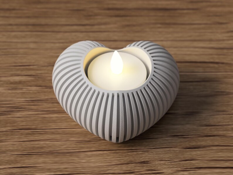 May include: A heart-shaped candle holder with a white, ribbed design. The holder contains a lit, white candle. The candle holder is made of a light gray material and sits on a wooden surface.