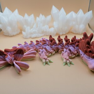 May include: An assortment of fantasy-themed sculptures in shades of white, pink, and brown. The collection includes crystal formations and a stylized dragon with a detailed head and body. The pieces are arranged on a light-colored surface.
