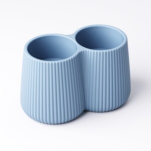 May include: A light blue, two-compartment container with a ribbed exterior. The container has a rounded base and two circular openings at the top. The design is simple and modern, suitable for holding items like pens or makeup brushes.