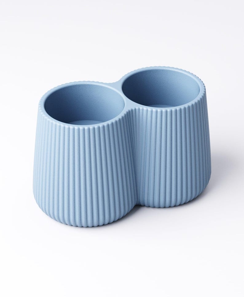 May include: A light blue, two-compartment container with a ribbed exterior. The container has a rounded base and two circular openings at the top. The design is simple and modern, suitable for holding items like pens or makeup brushes.