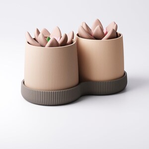 May include: Two beige, ribbed cylindrical planters with a dark grey base, each holding a faux succulent arrangement. The succulent petals are a soft pink with a touch of green. The planters are on a white background.