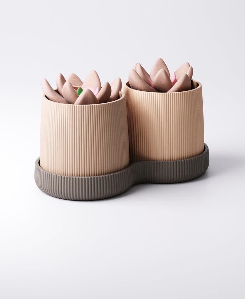 May include: Two beige, ribbed cylindrical planters with a dark grey base, each holding a faux succulent arrangement. The succulent petals are a soft pink with a touch of green. The planters are on a white background.
