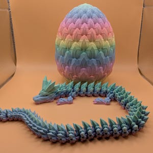 May include: A 3D-printed dragon and egg set. The dragon is articulated and blue-green, with a spiky spine. The egg is a pastel rainbow of pink, yellow, blue, and green. The set is displayed on an orange surface.