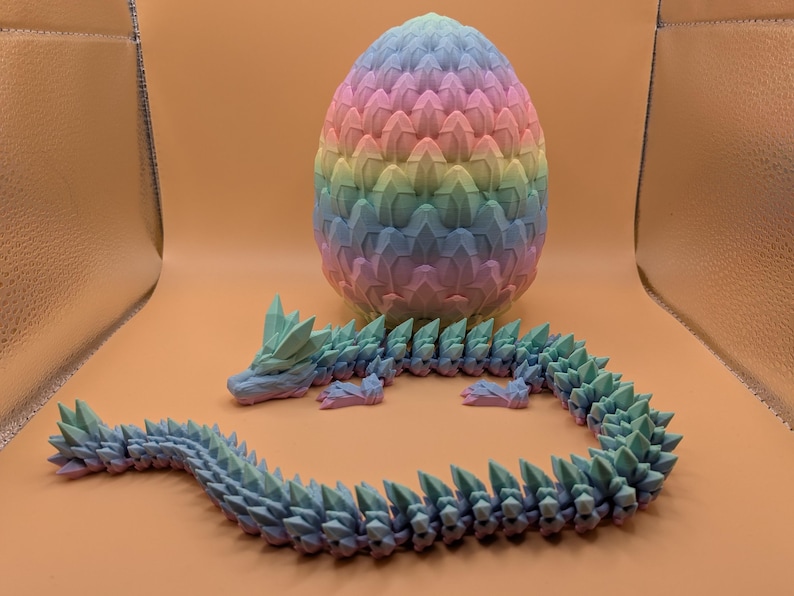 May include: A 3D-printed dragon and egg set. The dragon is articulated and blue-green, with a spiky spine. The egg is a pastel rainbow of pink, yellow, blue, and green. The set is displayed on an orange surface.