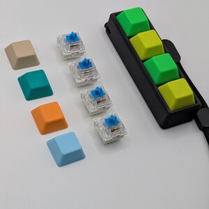 May include: A collection of colorful keyboard keycaps and switches. Keycaps are in beige, teal, orange, and light blue. Four switches are visible, with blue stems. A black holder with four green and yellow keycaps is also present.