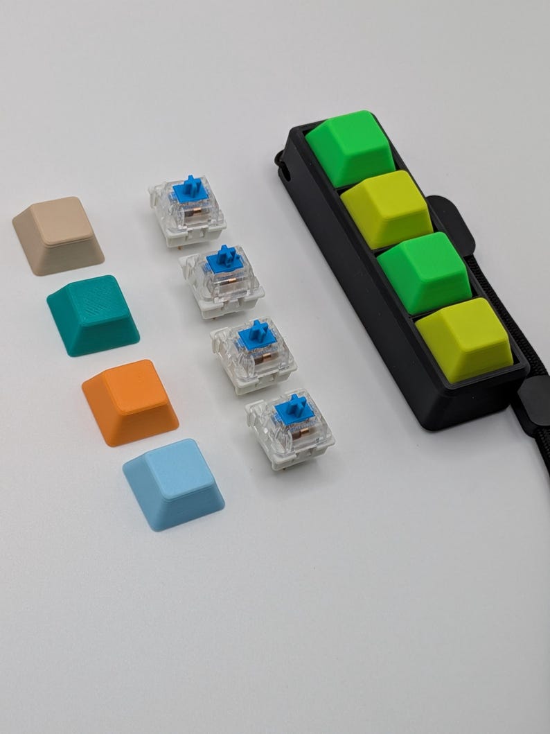 May include: A collection of colorful keyboard keycaps and switches. Keycaps are in beige, teal, orange, and light blue. Four switches are visible, with blue stems. A black holder with four green and yellow keycaps is also present.