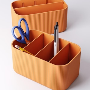 May include: Two orange desk tidies with vertical ribbed sides. One tidy contains a pair of blue scissors, a yellow pencil, and a silver pen. The other tidy is empty. The tidies are on a white surface.
