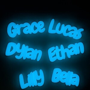 May include: Six names, Grace, Lucas, Dylan, Ethan, Lilly, and Bella, glow with a bright blue light against a dark backdrop. The names are written in a rounded, friendly font, giving a luminous appearance.
