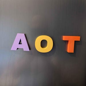 May include: Three colorful letters spelling "AOT" are displayed against a dark gray background. The letter "A" is purple, "O" is yellow, and "T" is orange. The letters appear to be made of a solid material.