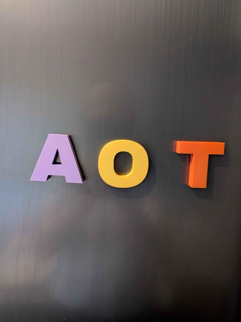 May include: Three colorful letters spelling "AOT" are displayed against a dark gray background. The letter "A" is purple, "O" is yellow, and "T" is orange. The letters appear to be made of a solid material.