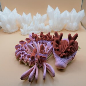 May include: The image showcases several 3D-printed sculptures. The foreground features a detailed, multi-colored dragon head and body in shades of pink and brown. Behind it are several white, crystalline structures, resembling clusters of crystals.