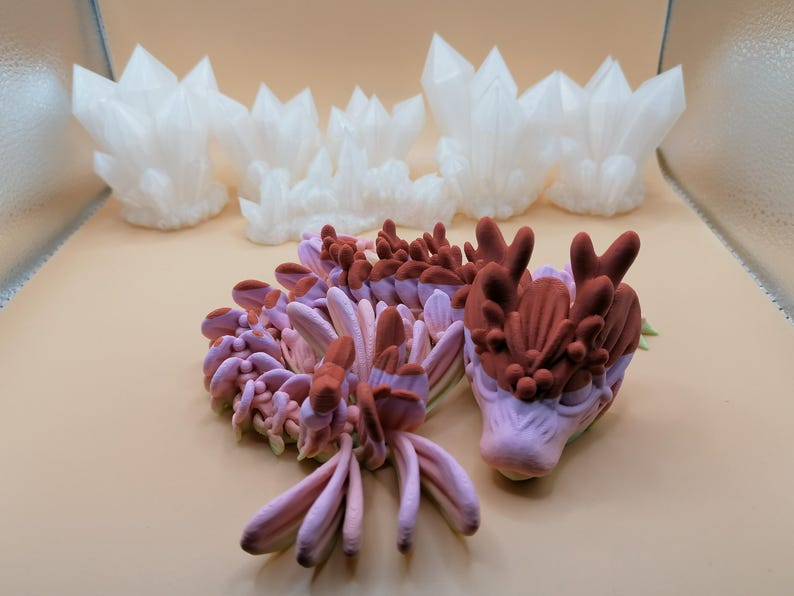 May include: The image showcases several 3D-printed sculptures. The foreground features a detailed, multi-colored dragon head and body in shades of pink and brown. Behind it are several white, crystalline structures, resembling clusters of crystals.