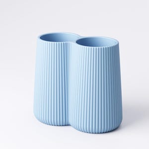 May include: A light blue, double-chambered vase with vertical ribbed detailing. The vase has a rounded base and a smooth rim. The two chambers are connected, creating a unique, modern design. The vase is made of a solid material and is suitable for home decor.